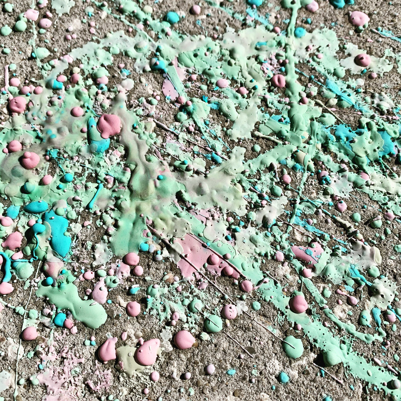This is a picture of several different colors of paint that has dried over a patch of gravel, most likely the remnants of a painting project. 