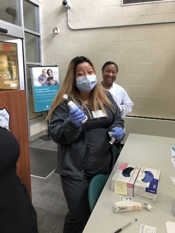 A healthcare worker wearing sterile gloves and a facemask holding a thermometer. Another person standing behind them without a facemask smiling. Both standing in front of a table with sterile gloves and another thermometer on top of it.  