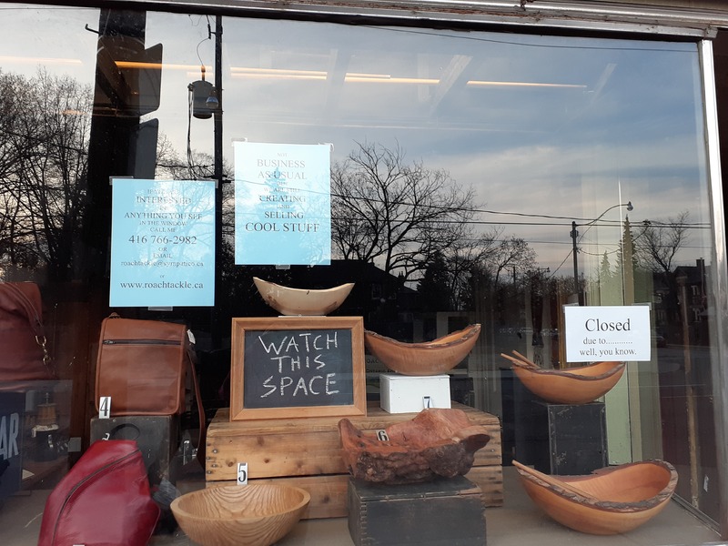 This is a picture of a storefront window, which has several bags and wooden bowls on display for sale. Signs on the window read "Not business as usual, but we are still creating and selling cool stuff", "watch this space", "Closed due to.... well you know.". Another sign implores the reader to call the owner if they are interested in buying anything on display. 