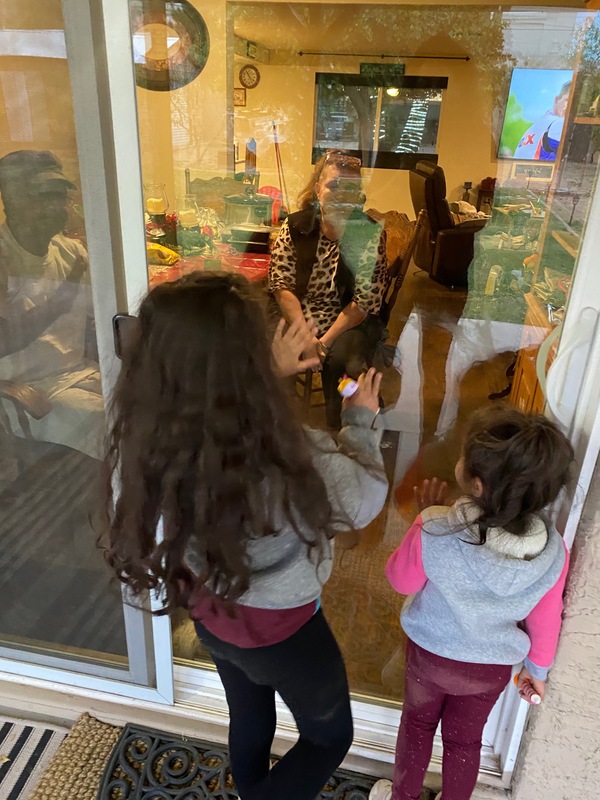 This is a picture taken of two girls staring through the sliding glass door of a home. On the other side, two older people wave at them. Both girls are dressed for winter weather, in grey and maroon clothes. 