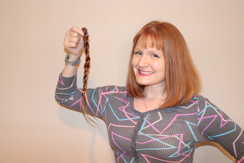 This is a picture of a woman smiling and posing with hair she has just cut from her head in the form of a braid. 