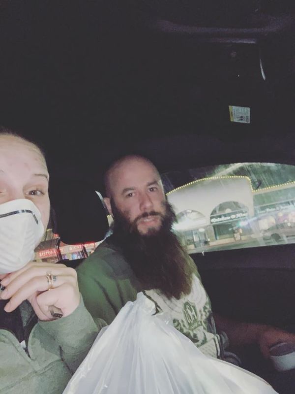 Two people are sitting in a car. The person on the left is wearing a face mask and has a green pullover hoodie on with food in a plastic bag in their lap. The person on the right has a long beard and is wearing a green shirt and also has a styrofoam drink in their hand. 