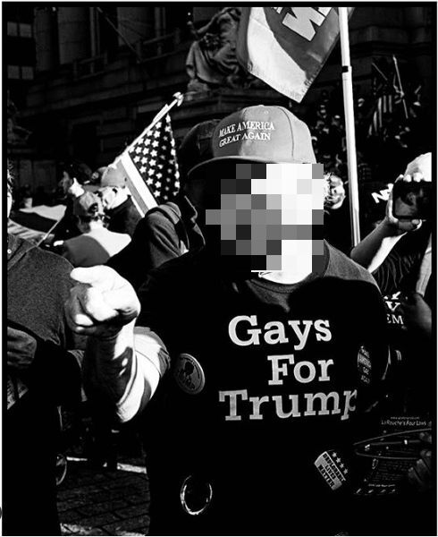 A man wearing a 'Make America Great Again' hat and a 'Gays For Trump' shirt points off camera at a Trump rally in New York City. His face is blurred. 