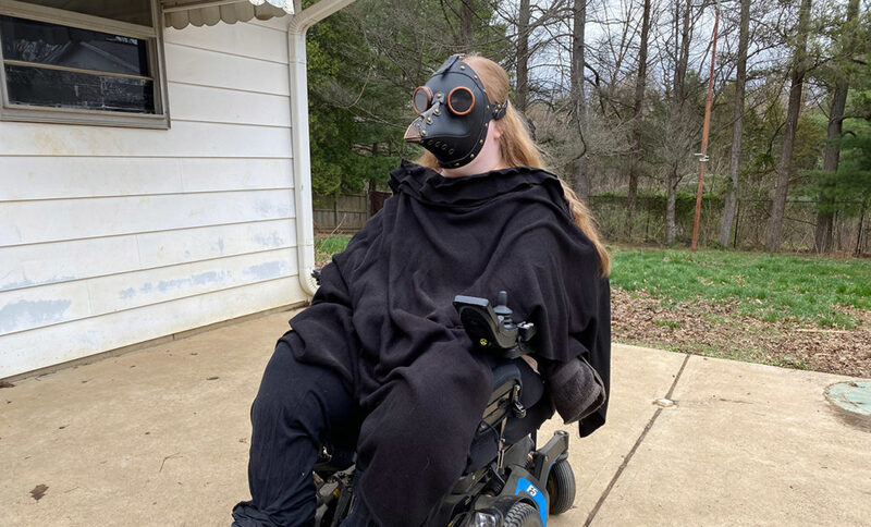 This is a picture of a woman in a motorized wheelchair, who is wearing black clothing and a mask that resembles those worn by Plague doctors during the Black Plague. 