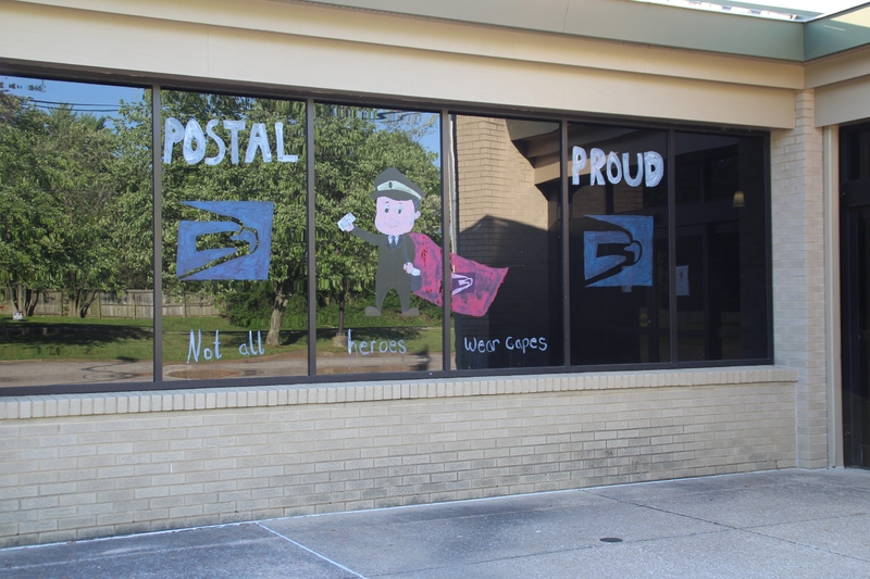 Window Writing Reading "Postal Proud. Not all Heroes Wear Capes" along with a drawing of a postal service man.