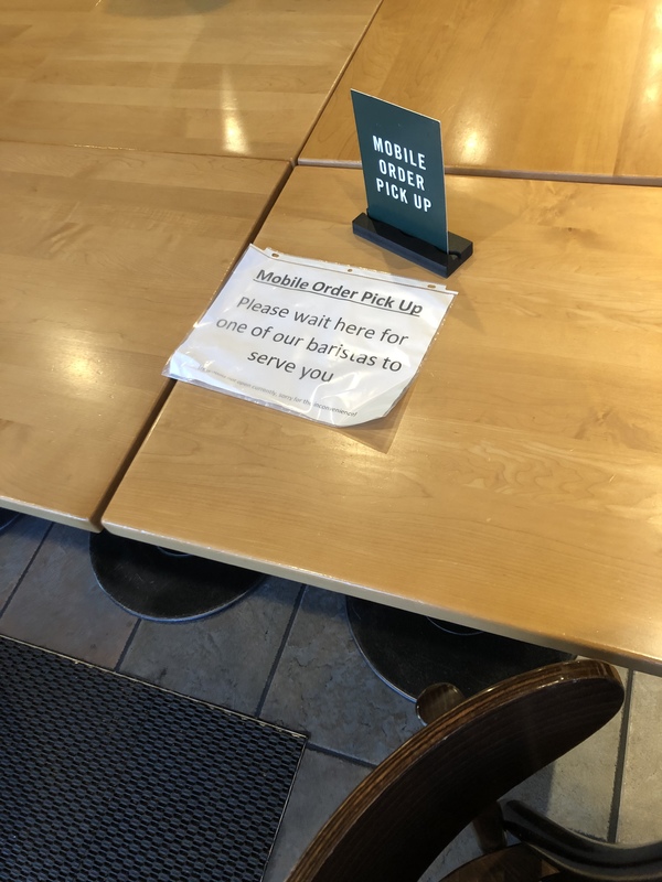 A table with two signs: a green one upright with text, "MOBILE ORDER PICK UP" and the other a paper in a sheet protector laying on the table in front of the green one with text, "Mobile Order Pick Up: Please wait here for one of our baristas to serve you"
