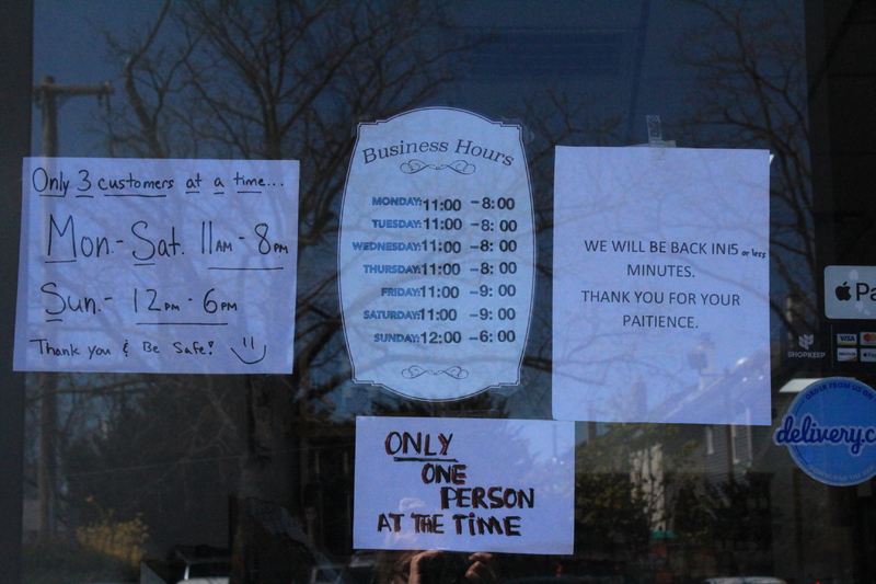 A business with multiple signs on the doors, one sign has amended business hours and says "three customers at a time" while another sign says "only one person at a time," the final sign is informing patrons that the business will reopen in fifteen minutes.