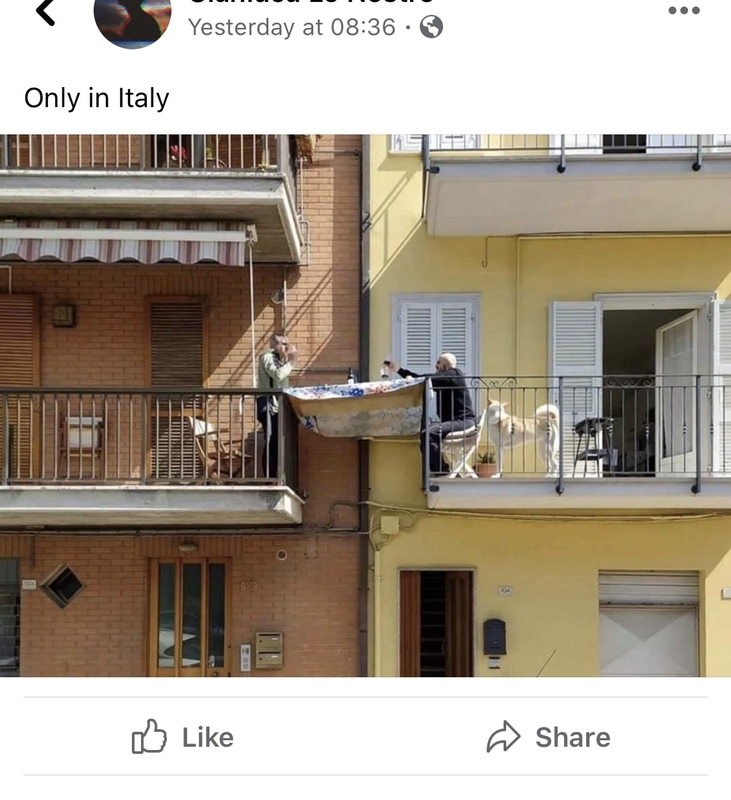 A screenshot of a post on Facebook where two neighbors share a glass of wine over a wooden table connecting their two balconies. 
