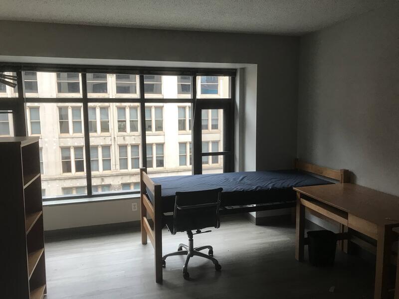 This is a picture taken of a mostly empty dorm room with a large bay window in the background overlooking the street. 