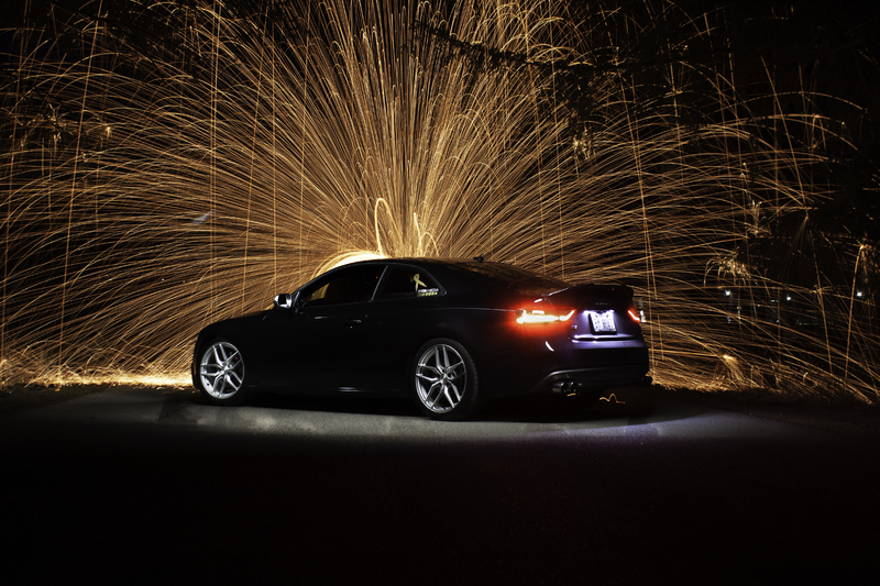 This is a picture of a sedan at night with fireworks going off directly behind it. 