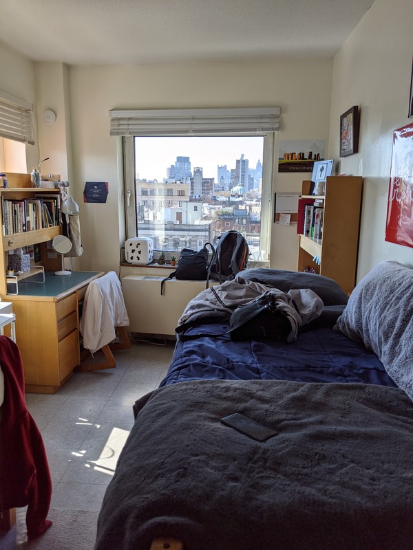 A white dorm room with tile flooring. The room has a window looking out at buildings that continue off into the distance. To the left of the room has a wooden desk that has a bookcase above it that is filled with books. On the right of the room has a bed that is leading out of frame, the bed has a navy bedspread with a grey folded blanket on it. In front of the bed in the corner of the room is another wooden bookcase filled with books. 