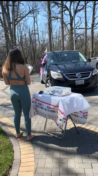 This is a picture of a young woman standing next to a table laden with desserts, with her back to the camera, staring at a new car. The table cloth reads "happy birthday". 