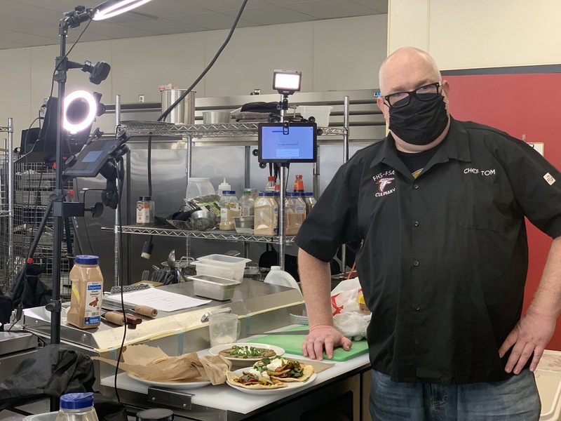 This is a picture of a bald man standing inside of a kitchen next to several plates of food he made. The man is wearing glasses, a face mask, jeans, and a shirt that reads "chef tom" in the right hand corner. A camera and portable lighting is set up in order to take pictures of the food. 
