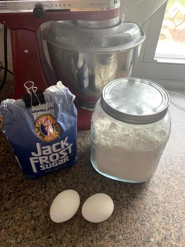 A red Artisan Kitchen Aid, a mason jar with a silver lid filled with white flour, two white eggs, and a blue Jack Frost granulated pure cane sugar bag on a speckled countertop surface. 