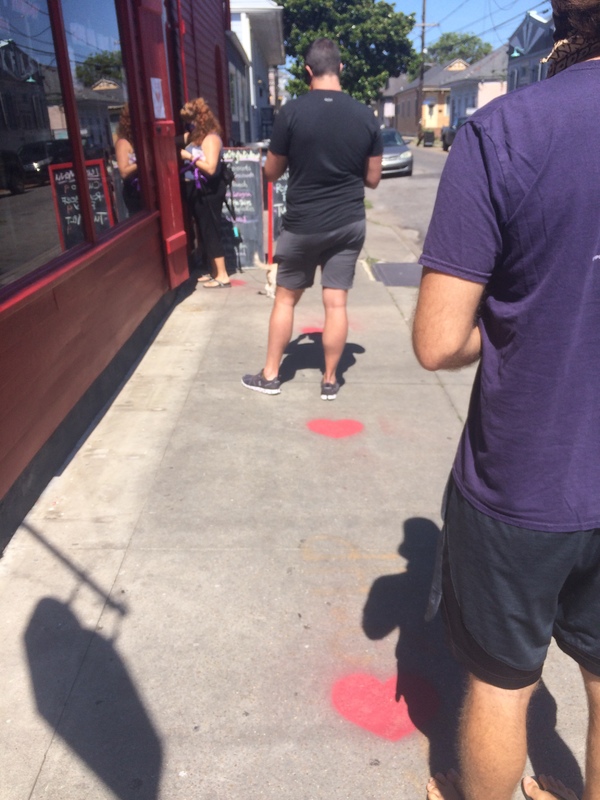 Customers lined up outside a bakery, waiting by heart shaped marks spray painted on the sidewalk that are six feet apart. 