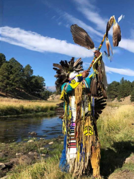 This is a picture of an indigenous man dressed in ceremonial clothing performing a type of traditional ceremony by a creek. 