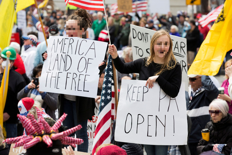 A crowd of white people protesting with their faces blurred. Two signs read "America Land of the Free" and "Keep PA open".