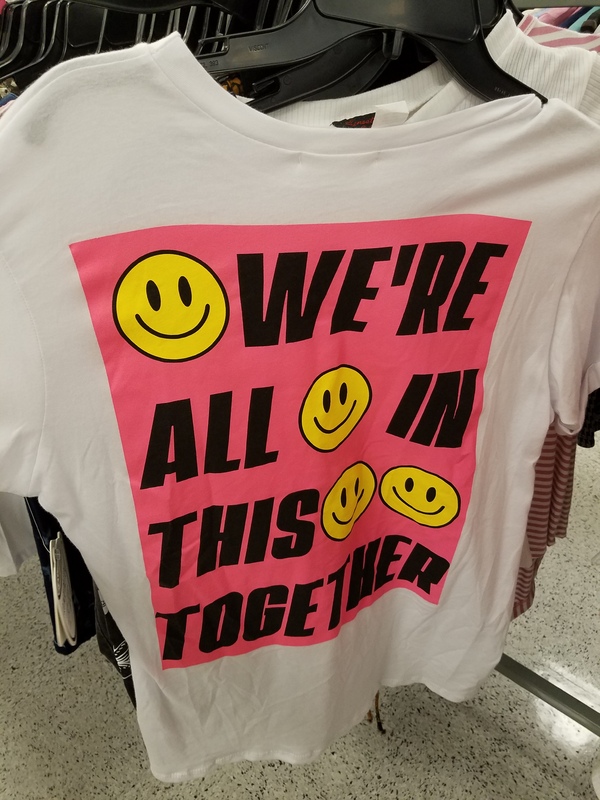This is a picture of a white and pink shirt covered with yellow smiley faces that says "We're all in this together" on it. 