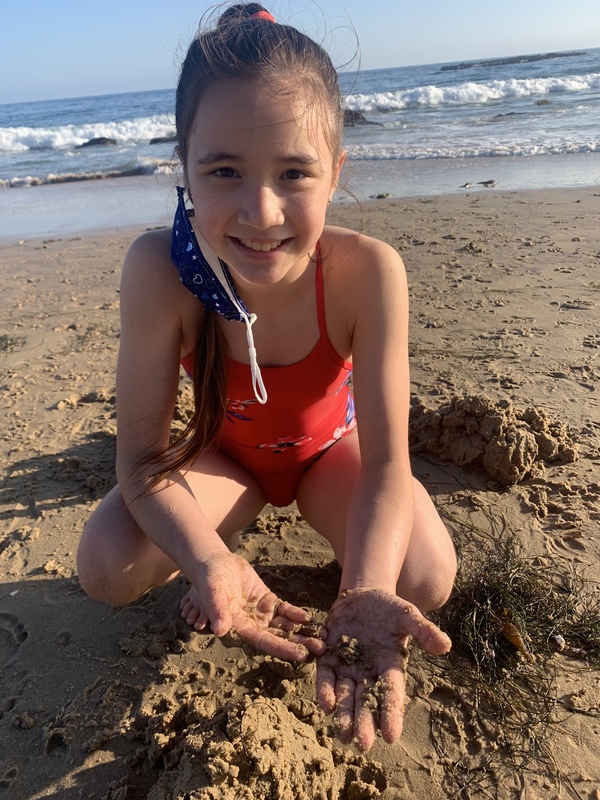 This is a picture of a young girl in a red swimsuit playing in the sand at the beach, and smiling into the camera. A face mask hangs off of one of her ears. 