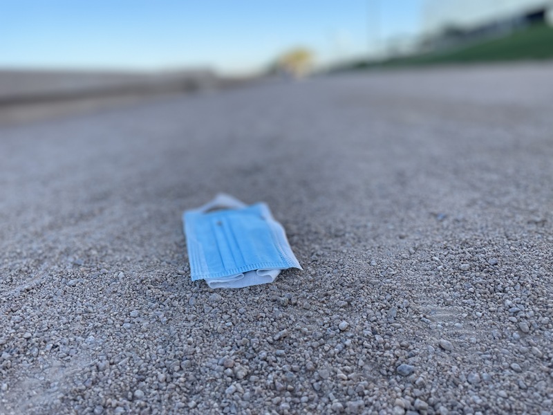 This is a picture of a blue face mask that has been discarded on a gravel and dirt path. The rest of the background is out of focus. 