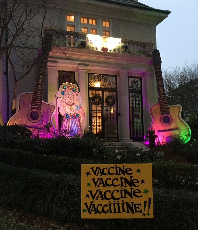 This is a picture of a house decorated with several COVID-19 related objects. A yellow sign stuck in the lawn out front reads "Vaccine, Vaccine, Vaccine, Vacciiiine!" Two giant acoustic guitars also flank the entrance to the home. 