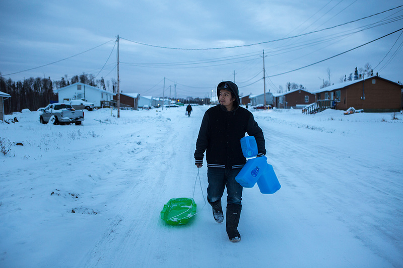 This is a picture of a man in dark winter clothing dragging a sled in the snow behind him, and carrying three jugs. 