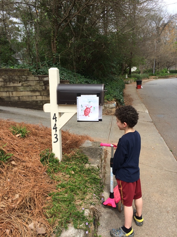 A kid is standing near a white mailbox in a neighborhood. The kid is wearing a navy long sleeve shirt with red shorts. The child is holding a pink and white scooter with four wheels on the bottom. Taped on the mailbox is a drawn picture of the Coronavirus. 