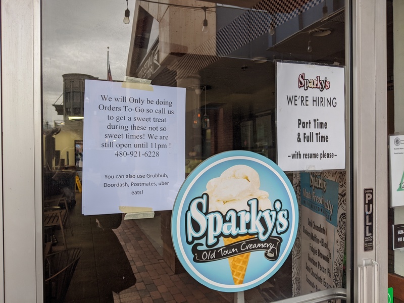 A glass front door leading into Sparky's Old Town Creamery has paper signs taped to the inside of the glass. The sign on the left says: We will Only be doing Orders To-Go so call us to get a sweet treat during these not so sweet times! We are still open until 11pm ! You can also use Grubhub, Doordash, Postmates, or uber eats! The sign on the right says: WE'RE HIRING Part Time & Full Time -with resume please-.