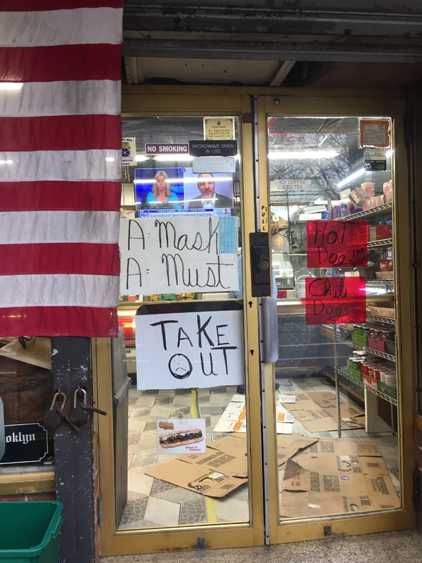 Store front double glass door with 2 white signs on the left with text, "A MASK A MUST" with a face mask by the word mask and "TAKE OUT." The right side has 2 red signs with text, "HOT DOG 1.00 TOPPINGS 1.50" and "CHILI DOGS 2.00"