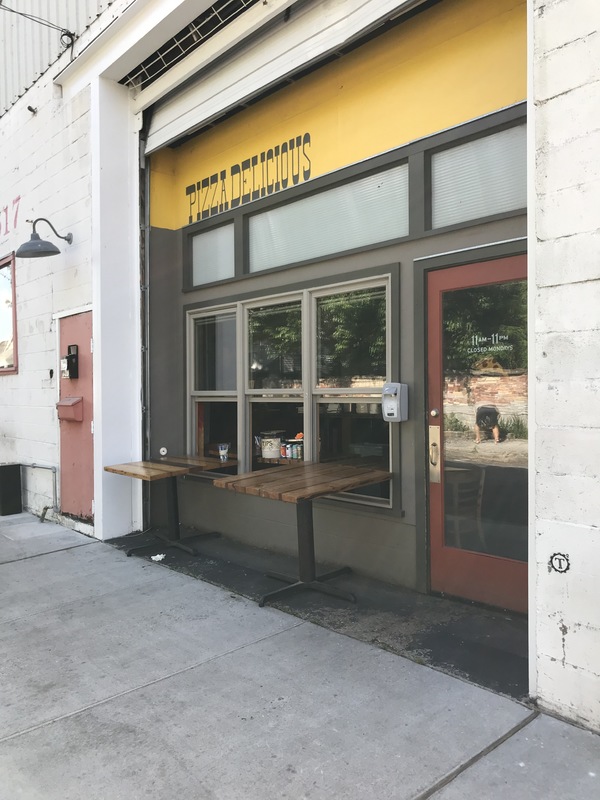 A dark grey and yellow building that says: PIZZA DELICIOUS across the top. The windows on the front of the building have wooden tables hanging out of them.