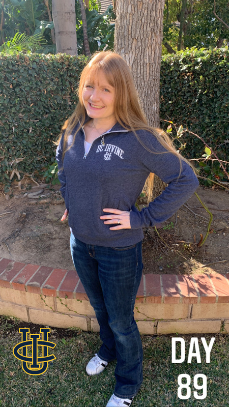 This is a picture of a girl wearing a UC Irvine sweatshirt and jeans smiling in front of a planting area with trees in the background. A UC Irvine logo is present in the left hand corner of the photo. 