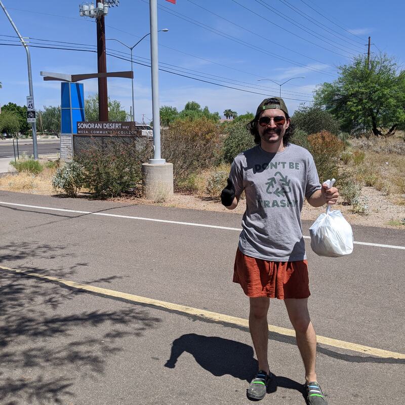 This is a picture of a man standing in a street smiling for the camera, while carrying a plastic grocery bag in one had, and flashing a thumbs up with the other. He is wearing sunglasses, shorts, and a shirt that reads "Don't be trash" on it with a recycling symbol in the center. 