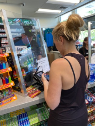 A person in a black tank top and blonde hair paying at the counter. Clear divider between employee and customer. 