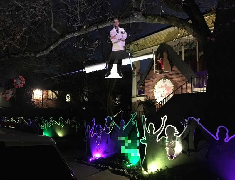 Exterior of home in New Orleans at night, lit with purple, green, and yellow lights.  Cardboard cut outs of people raising hands and dancing are on the ground, and hanging above, suspended from a tree, is a cardboard cut out of a doctor. 