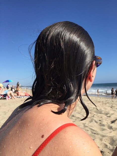 This is a picture taken from behind of a woman wearing sunglasses who just got out of the water staring out at the ocean. Her hair is wet, and she appears to be wearing a red swimsuit. 