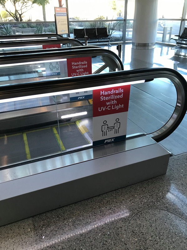 This is a picture of an escalator which has signs on it reading: "Handrails Sterilized With UV-C Light". 