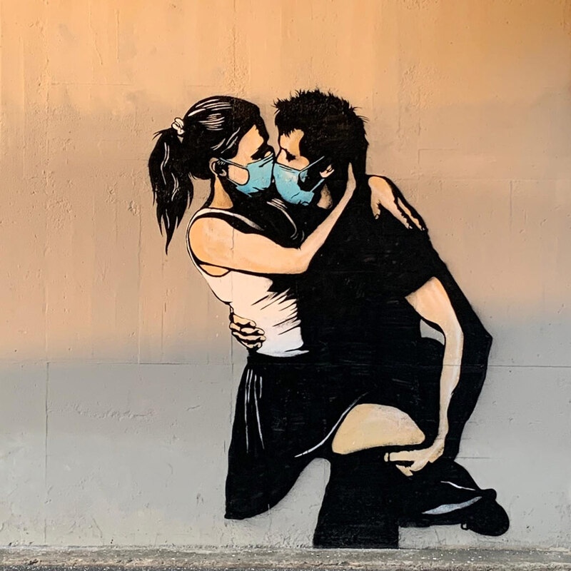 This is a picture of a mural on a wall which depicts a man holding a woman, who has her arms around his neck in a kissing pose. Both are shown wearing face masks. 