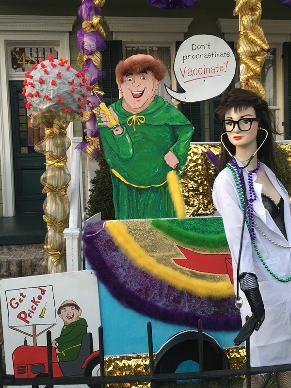 This is a picture of a series of decorations which encourage the viewer to make sure they get vaccinated. A character that resembles a monk clothed in a green robe is illustrated holding a needle and saying "Don't procrastinate, vaccinate!". Another mannequin dressed as a female doctor or nurse is next to this monk, and an object made to look like a COVID-19 particle stands on a pillar next to them. 