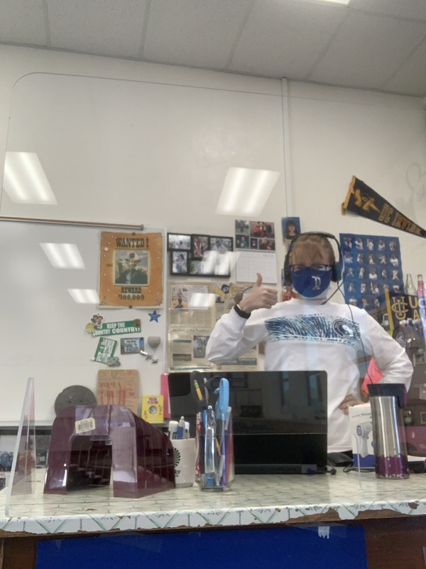 This is a picture of a woman in a white sweatshirt, wearing a face mask, glasses, and headphones. This picture appears to be taken in a classroom, with a whiteboard and other classroom decorations in the background. 