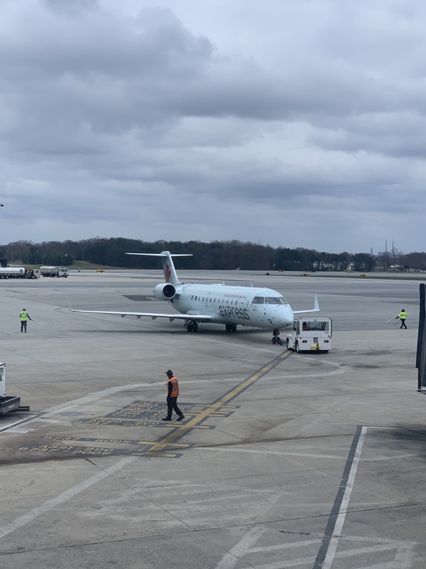 A white plane is parked outside. There are various people wearing orange and yellow safety vests walking around the plane. 