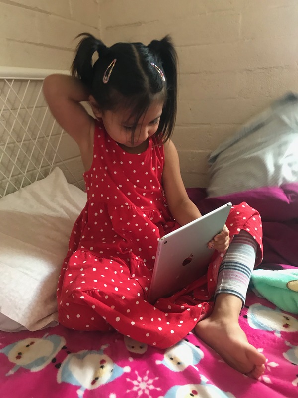 A girl with pigtails in a white and red polka dot dress is sitting on a bed with an iPad sitting in her lap. 