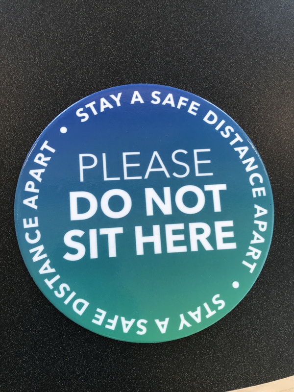 This is a picture of a blue sticker which reads "Stay a safe distance apart: please do not sit here"