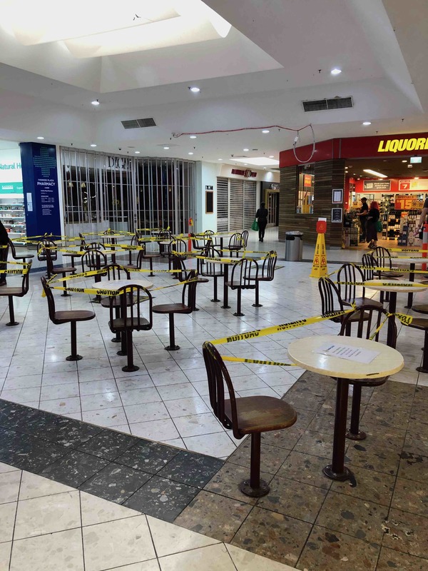 Inside of a shopping mall has caution yellow tape wrapped around seats and tables to block them off from people being able to use them. 