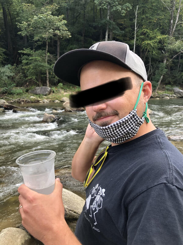 Image of a man sitting by a stream with a drink wearing a face mask over his chin. The subjects face is blurred. 