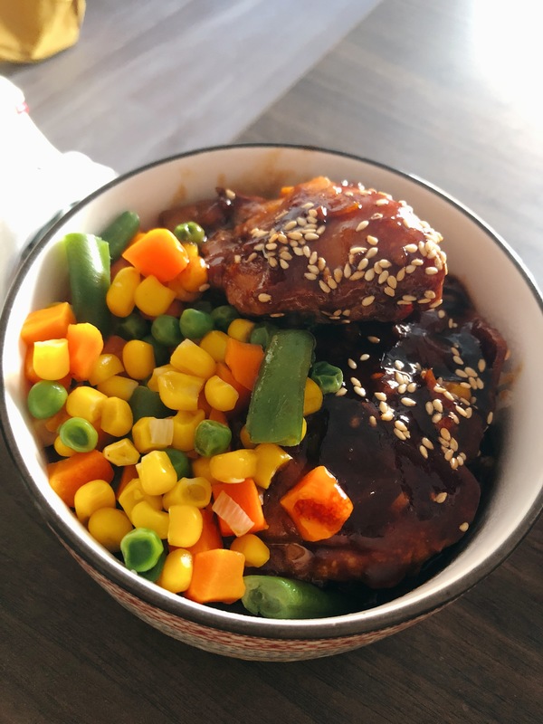 A bowl with meat covered in sauce with sesame seeds on top with vegetables on the side.