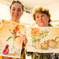 This is a picture taken of two women holding paintings of flowers in vases. 