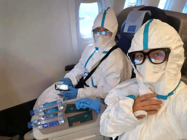 Two people are sitting on a plane with hazmat suits on posing for a selfie. 