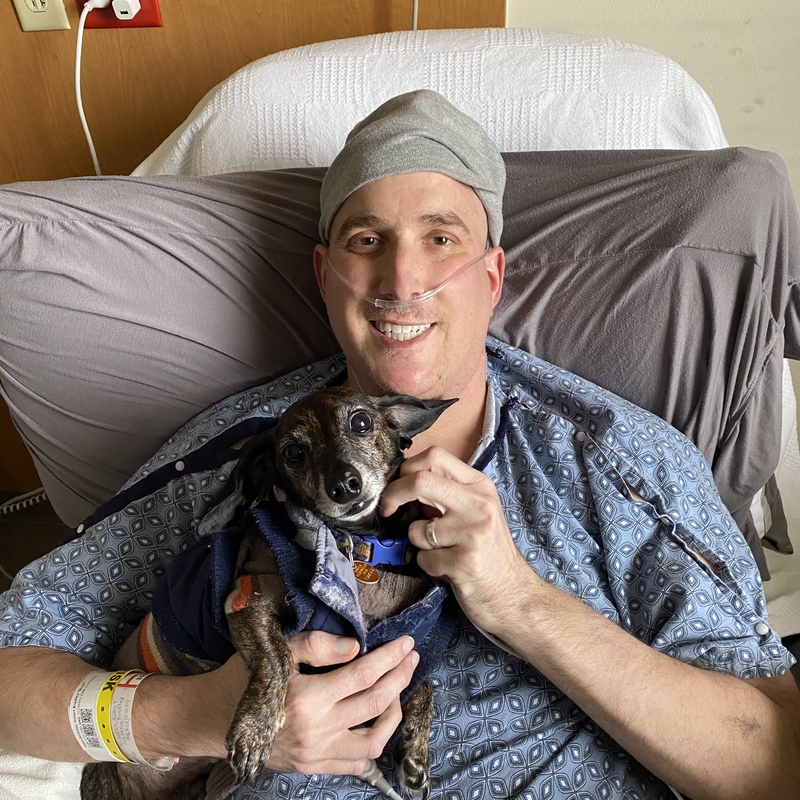 This is a picture of a man wearing a hospital gown, sitting in a bed at a hospital holding a dog and smiling. 