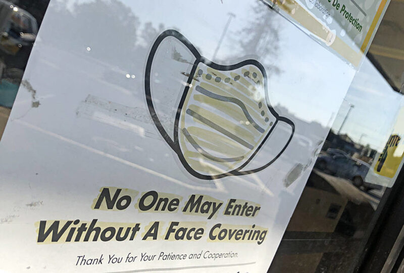 This is a picture of a sign posted on the inside of a window, on which the image of a face mask can be seen along with the words "No One May Enter Without A Face Covering- Thank You for Your Patience and Cooperation. "