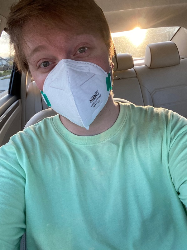 A selfie of a man wearing a green shirt and a surgical mask sitting in his car.